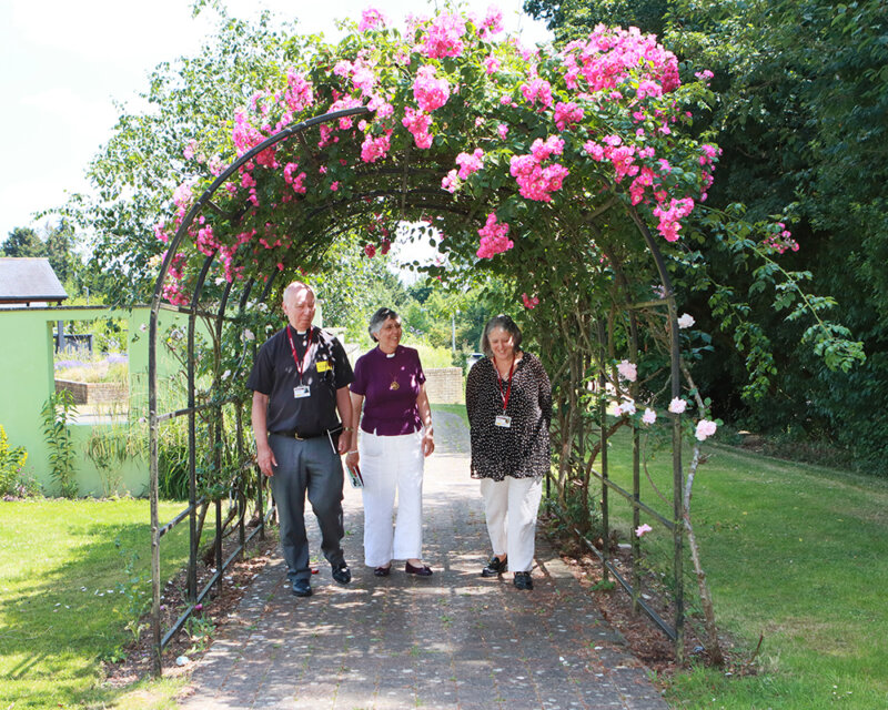Rev Andrew Merchant, Dr Guli Francis Dehqani, Michelle Kabia walk in the grounds of Farleigh Hospice under a rose tree archway
