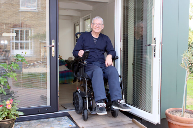 Richard is shown smiling while sitting in his electric wheelchair in the doorway of his new home