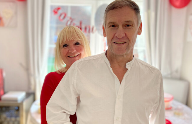 Debbie stands behind Kevin giving him a hug at a party with balloons and decorations in the background. Both are smiling. hugging Kevin from behind