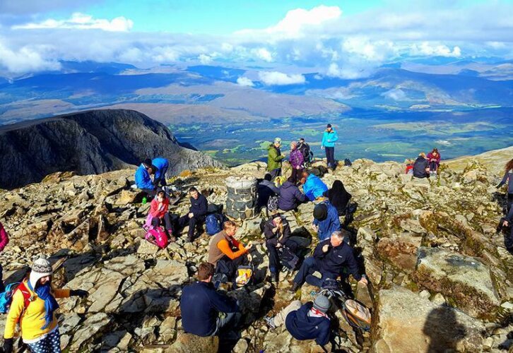 A diverse group of individuals atop a mountain, smiling and taking in the stunning scenery below.