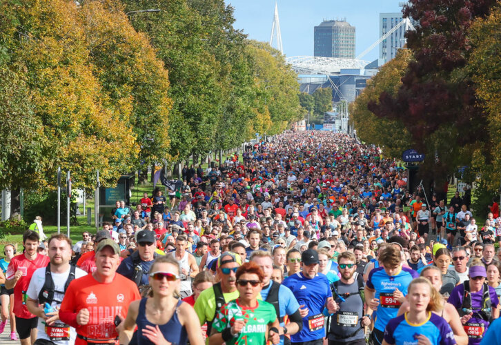 A large group of runners participates in a marathon on a tree-lined city street, with buildings visible in the background under a clear sky.