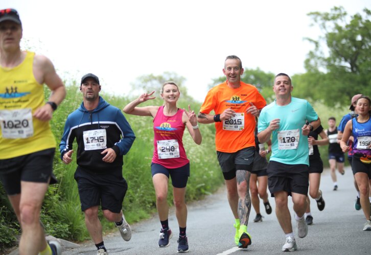 A group of marathon runners races along a rural road bordered by tall green foliage, featuring diverse athletes in colorful running gear and numbered race bibs. A smiling woman in a pink athletic tank top makes a peace sign while running beside others wearing bright orange, teal, yellow, and blue shirts. The scene captures outdoor endurance running, competitive racing energy, and a community fun run atmosphere under an overcast sky.