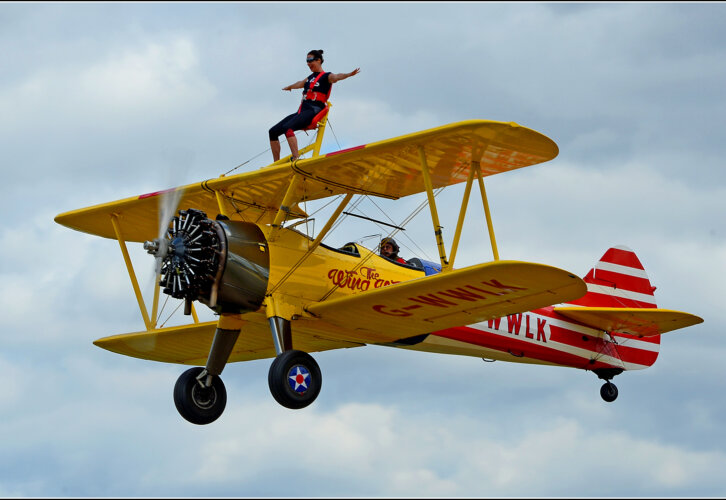A person stands on top of the upper wing of a yellow biplane in flight, while another person pilots the plane against a cloudy sky.