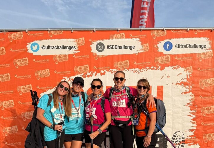 Five ladies standing together at the finish line wearing bright neon coloured branded Farleigh Hospice tshirts with sunglasses and bagpacks with huge smiles on their faces.