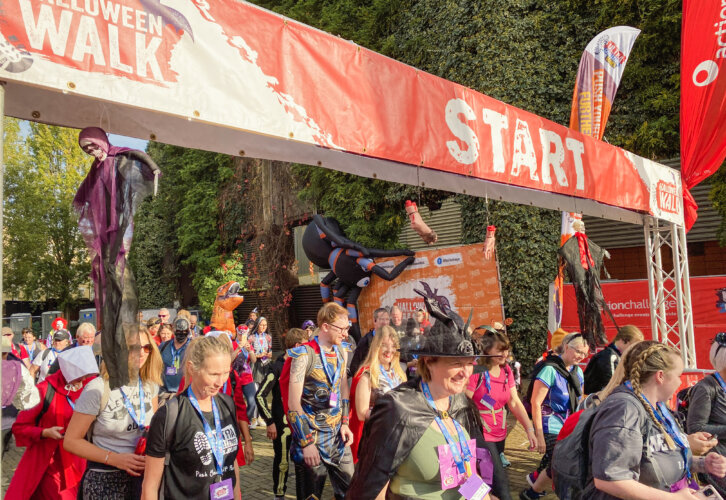 Costumed participants begin a festive outdoor event under a "Halloween Walk Start" banner. They wear diverse Halloween outfits, exuding excitement.