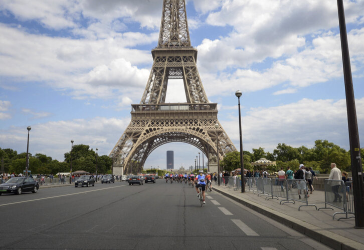 A picture of the Eiffel Tower in Paris, France surrounded by clouds in the sky and bike riders riding along side cars driving towards the tower.
