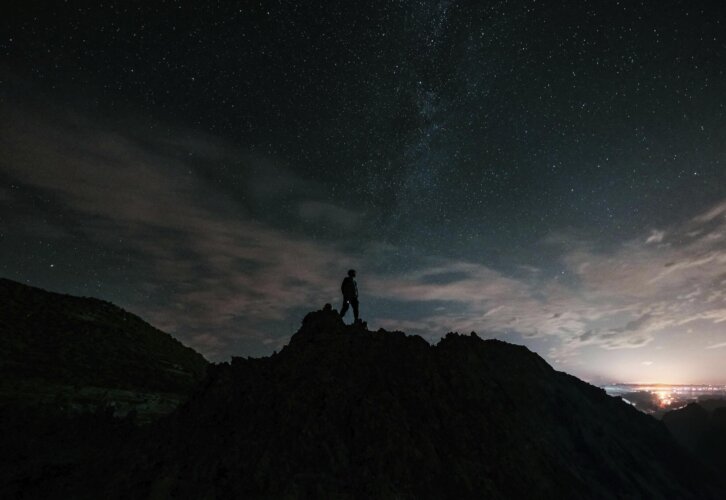 A man stands on a mountain summit under a night sky, illuminated by stars, capturing a moment of reflection and awe.