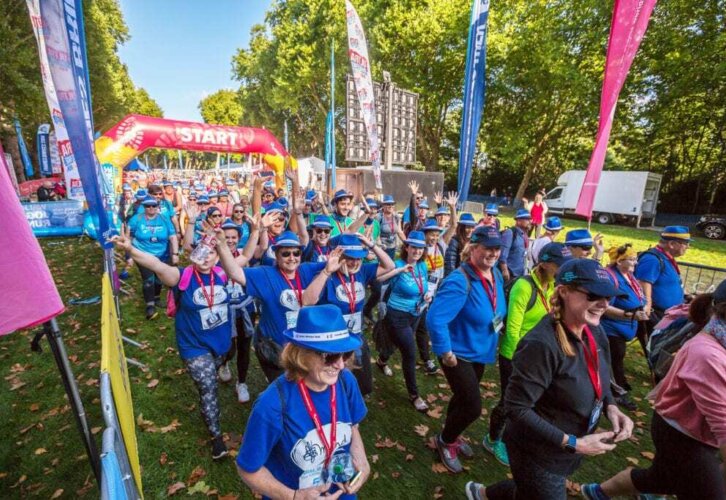 A joyful crowd of people in colorful attire, wearing blue hats and red medals, gathers at a race start line under a bright sky with trees nearby.