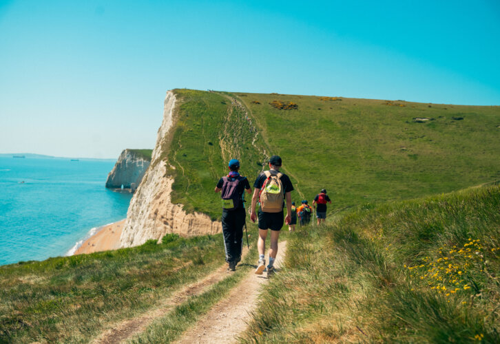A scenic mountain view by the seaside with green grass and a bright blue sky, with a far away shot of people walking on a trail with hats and backpacks on.