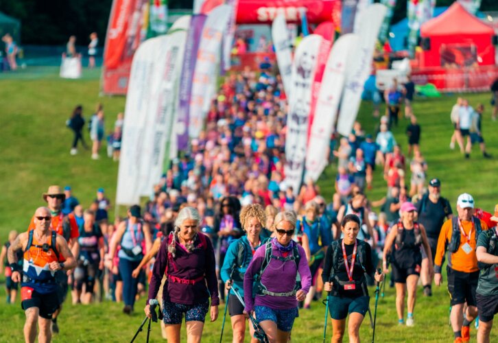 A big group of people with walking gear and backpacks walking along a grassy field with branded flags surrounding them.