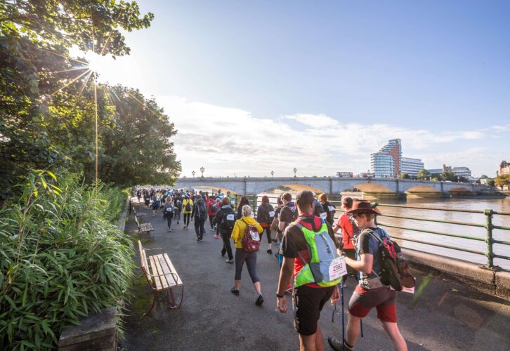 Several individuals walk together on a path beside the Thames river, surrounded by the sun, greenery and the sound of flowing water.