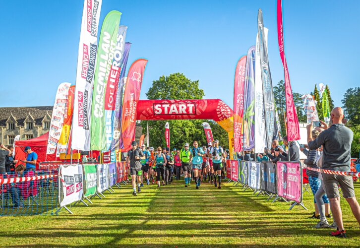A diverse group of people strides confidently through a colourful, sunny race, showcasing determination and camaraderie. With people on the sides taking photos of participants.